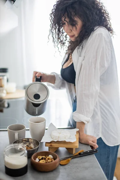 Uśmiechnięta kręcona kobieta przygotowuje napój ziołowy wlewając gorącą wodę z czajnika do filiżanki z torebką herbaty — Zdjęcie stockowe
