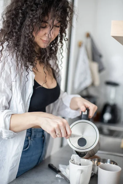 Uśmiechnięta kręcona kobieta przygotowuje napój ziołowy wlewając gorącą wodę z czajnika do filiżanki z torebką herbaty — Zdjęcie stockowe