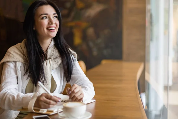 Felice Asiatica giovane donna seduta al caffè con tazza di caffè prendere appunti godendo di pausa — Foto Stock