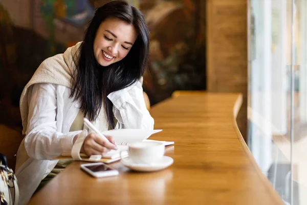 Feliz joven asiática sentada en la cafetería con taza de café tomando notas disfrutando de un descanso —  Fotos de Stock