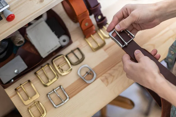 Mãos de close-up de bronzeador profissional escolhendo fivela de cinto na mesa de madeira na oficina de couro — Fotografia de Stock