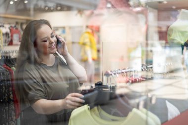 Mutlu ve boyut olarak güzel bir kadın mağazadan kıyafet seçiyor ve akıllı telefonlardan konuşuyor.