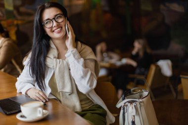 Güzel bir iş kadını, kafede dizüstü bilgisayar ve kahve fincanı ile akıllı telefondan konuşuyor.