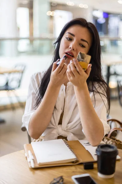 Hermosa joven mujer de negocios corregir el maquillaje con lápiz labial y espejo sentado en la mesa de la cafetería — Foto de Stock