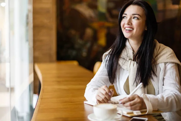 Feliz joven asiática sentada en la cafetería con taza de café tomando notas disfrutando de un descanso —  Fotos de Stock