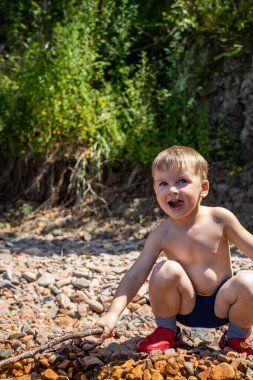 Çakıl taşlı sahilde oturan komik çocuk. Elinde doğal tahta sopayla deniz suyunda oynuyor. Eğleniyor.