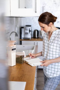 Gülümseyen genç iş kadını modern İskandinav mutfağında kağıt günlüğe not alıyor.