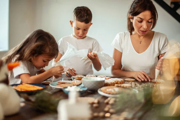 Mamma e bambini decorano il pan di zenzero di Natale a casa. Un ragazzo e una ragazza dipingono con cornetti con la glassa di zucchero sui biscotti. Decorazione di anno nuovo, rami di un albero di Natale. — Foto Stock