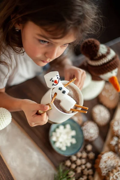 Little cute girl plays with knitted snowmen and eats gingerbread and drinks cocoa with marshmallows. — Stock Photo, Image