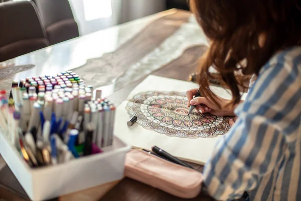 Jovem mulher bonita está sentada na cozinha da casa e desenhando uma mandala para colorir. Desenho zen. — Fotografia de Stock