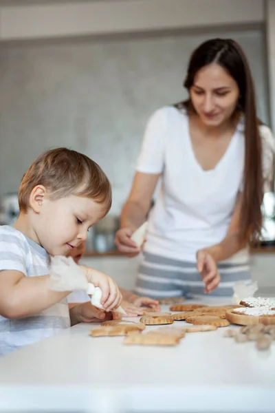 Mamma e bambini decorano il pan di zenzero di Natale a casa. Un ragazzo e una ragazza dipingono con cornetti con la glassa di zucchero sui biscotti. Decorazione di anno nuovo, rami di un albero di Natale. — Foto Stock