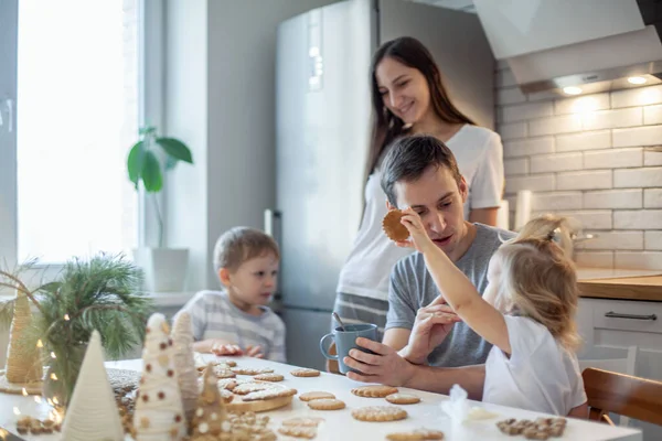 Dad, mom and children decorate Christmas gingerbread at home. A boy and a girl paint with cornets with sugar icing on cookies. New Years decor, branches of a Christmas tree. — Stock Photo, Image