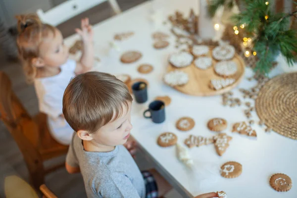 男の子と女の子は自宅でクリスマスのジンジャーブレッドを飾る。男の子と女の子がクッキーに砂糖アイシングでコーネットでペイントします。新年の装飾、クリスマスツリーの枝. — ストック写真