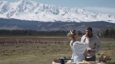 A young married couple on a picnic against a backdrop of snow-capped mountains. The woman feeds the man food from her hand. Family hiking in the mountains.