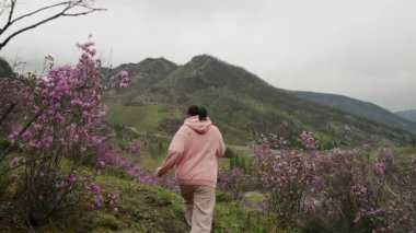 Two female tourists run up a mountain with flowering bushes, rejoicing and enjoying the beautiful views of the green mountains.
