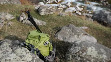 Dağ nehrinin yakınındaki kayalarda dinlenen yürüyüş direkleri olan yeşil bir sırt çantası. Seyahat, yürüyüş, macera, kamp ve aktif yaşam tarzı kavramı.