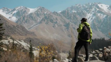 Yeşil ceketli ve sırt çantalı bir erkek yürüyüşçü kayalık arazide duruyor, görkemli, karlı dağ sıralarına bakıyor. Doğada macera ve özgürlük temalarını yakalamak. 