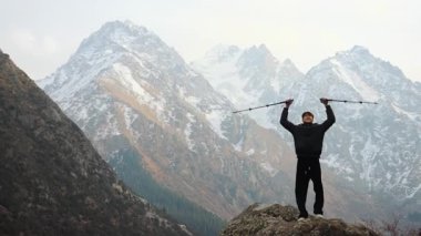 Genç bir adam muzaffer bir şekilde kayalık bir dağın zirvesinde dikiliyor, karla kaplı zirvelerin zeminine karşı tepe direkleri dikiyor. Başarı kavramı, özgürlük, macera, başarı.