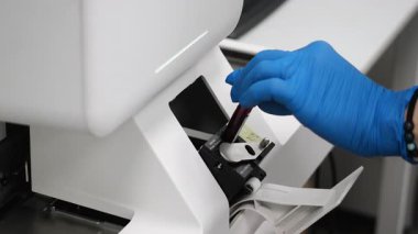 Mavi eldivenli bilim adamı, parlak steril ışık altında modern laboratuvarda otomatik klinik analiz cihazına kan testi tüpü yüklüyor..