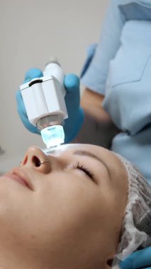 Modern bir estetik klinikte bir hastaya yüz LED cihazı tedavisi uygulayan bir kozmetolojist kapalı bir güzellik prosedürü sırasında profesyonel cilt bakım ekipmanı kullanıyor..