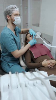 Diş hekimi, dişçi koltuğunda yatan bir hastaya diş tedavisi uyguluyor, profesyonel diş aletleri kullanıyor ve modern bir klinikte kapalı bir ameliyat sırasında koruyucu giysi giyiyor..