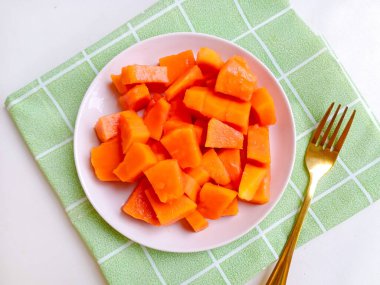 Beyaz bir tabakta altın çatalla servis edilen taze papaya küpleri. Sağlıklı gıda konsepti, temiz arka plan, doğal ışık, tropikal meyve düzlüğü