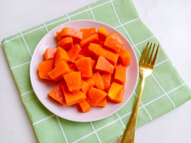 Beyaz bir tabakta altın çatalla servis edilen taze papaya küpleri. Sağlıklı gıda konsepti, temiz arka plan, doğal ışık, tropikal meyve düzlüğü