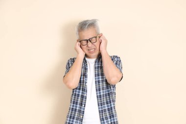 Gözlüklü, kulağı yakın, günlük ekose gömlek, şort ve spor ayakkabı giyen, bej arka planda izole Asyalı bir adamın portresi. Yaşlı erkek yaşam tarzı konsepti.