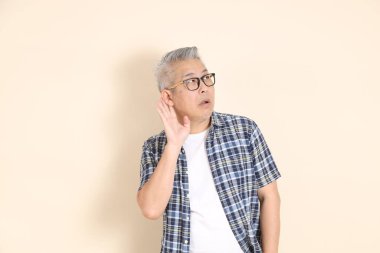 Gözlüklü, dinleyen ya da duyan, günlük ekose gömlek, şort ve spor ayakkabı giyen, bej arka planda izole Asyalı son sınıf öğrencisinin portresi. Yaşlı erkek yaşam tarzı konsepti.