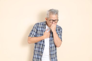 Gözlüklü, hasta Asyalı bir adamın portresi. Günlük ekose gömlek, şort ve spor ayakkabı giyiyor. Bej arka planda izole edilmiş. Yaşlı erkek yaşam tarzı konsepti.