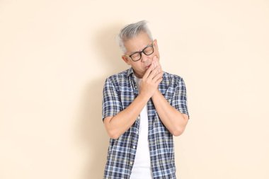 Gözlüklü, diş ağrısı çeken, günlük ekose gömlek, şort ve spor ayakkabı giyen bej arka planda izole Asyalı bir adamın portresi. Yaşlı erkek yaşam tarzı konsepti.