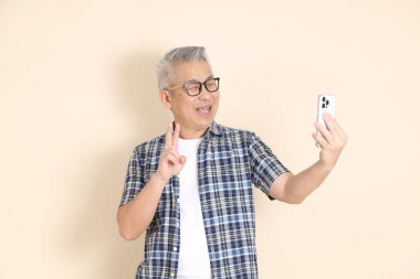 Gözlüklü, selfie çeken, fotoğraf çekmek için fotoğraf çeken, günlük ekose gömlek, şort ve spor ayakkabı giyen, bej arka planda izole edilmiş Asyalı son sınıf öğrencisinin portresi. Yaşlı erkek yaşam tarzı konsepti.