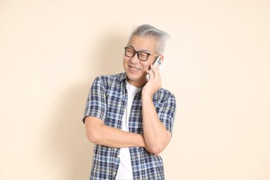 Gözlüklü, cep telefonu tutan, günlük ekose gömlek, şort ve spor ayakkabı giyen, bej arka planda izole Asyalı bir adamın portresi. Yaşlı erkek yaşam tarzı konsepti.