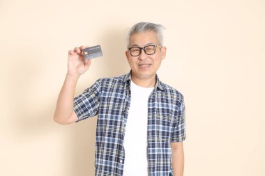 Gözlüklü bir Asyalı 'nın portresi, cep telefonlu kredi kartı, ekose gömlek, şort ve spor ayakkabı giyiyor, bej arka planda izole edilmiş. Yaşlı erkek yaşam tarzı konsepti.