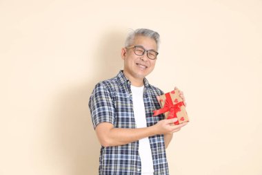 Gözlüklü, elinde hediye kutusu olan, günlük ekose gömlek, şort ve spor ayakkabı giyen, bej arka planda izole edilmiş Asyalı bir son sınıf öğrencisinin portresi. Yaşlı erkek yaşam tarzı konsepti.