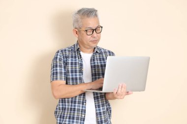 Gözlüklü, dizüstü bilgisayarı olan, günlük ekose gömlek, şort ve spor ayakkabı giyen, bej arka planda izole Asyalı bir adamın portresi. Yaşlı erkek yaşam tarzı konsepti.