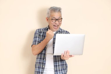 Gözlüklü, dizüstü bilgisayarı olan, günlük ekose gömlek, şort ve spor ayakkabı giyen, bej arka planda izole Asyalı bir adamın portresi. Yaşlı erkek yaşam tarzı konsepti.