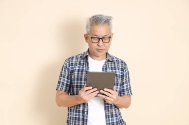 Gözlüklü, tablet tutan, günlük ekose gömlek, şort ve spor ayakkabı giyen, bej arka planda izole Asyalı yaşlı bir adamın portresi. Yaşlı erkek yaşam tarzı konsepti.