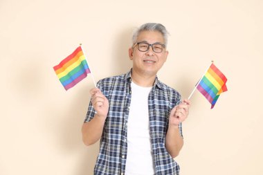 Gözlüklü, elinde bayraklar tutan, günlük ekose gömlek, şort ve spor ayakkabı giyen, bej arka planda izole Asyalı bir son sınıf öğrencisinin portresi. Yaşlı erkek yaşam tarzı konsepti.