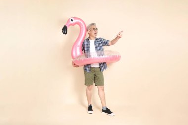 Gözlüklü, büyük pembe flamingo şişirilebilir yüzme yüzüğü tutan, bej arka planda izole günlük ekose gömlek giyen Asyalı son sınıf öğrencisi. Yaşlı erkek yaşam tarzı konsepti.