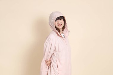 Pembe ultraviyole koruyucu kapüşonlu ceket giyen genç Asyalı kadın bej arka planda duruyor ve gülümsüyor..