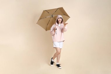 Pembe ultraviyole koruyucu kapüşonlu ceket giyen genç Asyalı bir kadın elinde bej arka plan, güneş koruması kıyafet konseptine karşı şemsiye tutuyor..