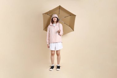 Pembe ultraviyole koruyucu kapüşonlu ceket giyen genç Asyalı bir kadın elinde bej arka plan, güneş koruması kıyafet konseptine karşı şemsiye tutuyor..