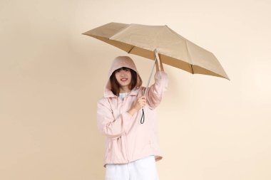 Pembe ultraviyole koruyucu kapüşonlu ceket giyen genç Asyalı bir kadın elinde bej arka plan, güneş koruması kıyafet konseptine karşı şemsiye tutuyor..