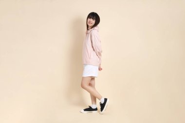 Pembe ultraviyole koruyucu kapüşonlu ceket giyen genç Asyalı kadın bej arka planda duruyor ve gülümsüyor..