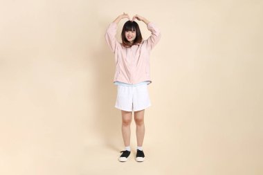 Pembe ultraviyole koruyucu kapüşonlu ceket giyen genç Asyalı bir kadın. Kalp şeklinde, mini kalp, bej arka planda aşk, güneş koruma giysisi konsepti..