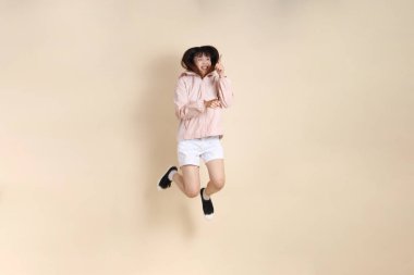 Pembe ultraviyole koruyucu kapüşonlu ceket giyen genç Asyalı bir kadın bej arka plana atlıyor, güneş korumalı giysi konseptine atlıyor..