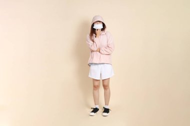 Yüzü maskeli pembe ultraviyole koruyucu kapüşonlu bir ceket giyen genç Asyalı bir kadın bej arka plan, güneş koruması kıyafet konseptine karşı hastayken....