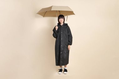 Siyah yağmurluk giyen ve elinde şemsiyesi olan genç bir Asyalı kadın bej bir arka planda tek başına duruyor. Yağmur mevsimi, hava durumu, sigorta kavramı.