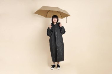 Siyah yağmurluk giyen ve elinde şemsiyesi olan genç bir Asyalı kadın bej bir arka planda tek başına duruyor. Yağmur mevsimi, hava durumu, sigorta kavramı.